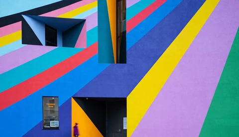 Colorful geometric mural with diagonal stripes in blue, pink, yellow, purple, and green, featuring black architectural elements and doorways.