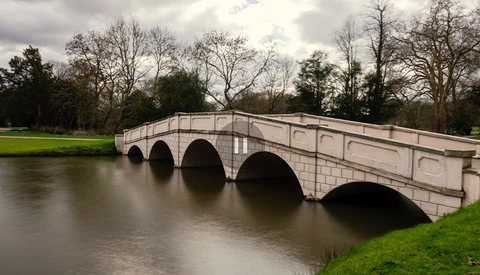 Stone arch bridge spanning a calm river with bare trees and green lawn in overcast daylight.