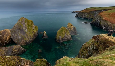 Moss-covered sea stacks and rocky outcrops in turquoise coastal waters beneath dramatic overcast skies.