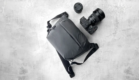 Flat lay of camera gear including DSLR body with attached lens, shoulder bag, and lens cap on concrete surface.
