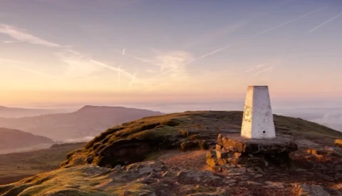White trig point beacon on a grassy hilltop at golden hour sunrise, with layered valleys and mountains in misty background.