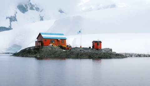 Orange and red buildings with blue roof on rocky island surrounded by icy water and snow-covered mountains.