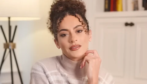 Woman with curly dark hair styled in a half-up bun, wearing a light pink ribbed sweater, posing with hand near face in a bright interior setting.