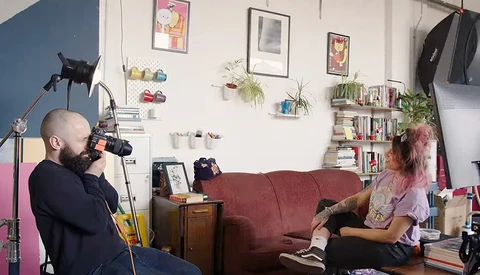 Filmmaker operating camera on tripod while interviewing subject seated on burgundy couch in home studio.