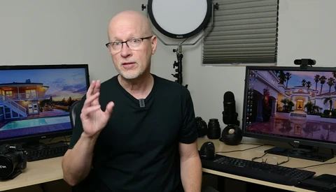 Man in black shirt gesturing while speaking in a studio with multiple monitors displaying architectural photography.