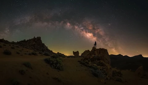 Silhouetted figure standing on a rocky outcrop beneath the Milky Way galaxy arcing across a starry night sky.