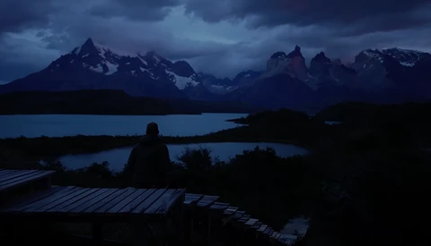 Silhouetted figure sitting on a wooden deck overlooking a mountain lake at dusk.