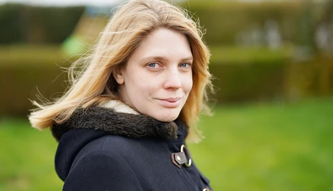 Woman with blonde hair looking over shoulder outdoors, wearing black jacket with fur collar.