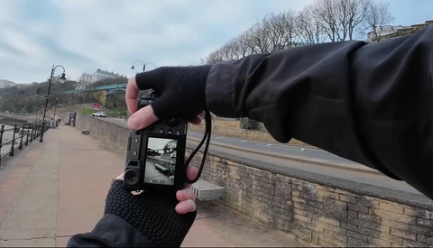 Photographer's hands holding a smartphone to frame a riverside promenade with bare trees and stone walls.
