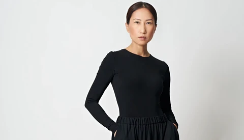 Woman in black long-sleeve top and gathered skirt standing against neutral background.
