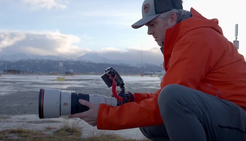 Photographer holding a telephoto lens with red hood near a frozen lake, mountains visible across water.