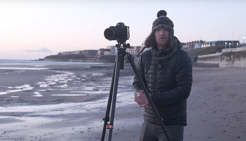 Photographer with camera on tripod on a beach at dusk, wearing winter clothing and beanie.
