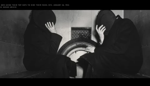 Two figures in dark clothing hide their faces with their hands while sitting near a large cylindrical metal object.