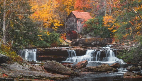 Historic gristmill with red roof beside cascading waterfall in autumn forest.