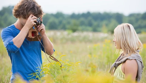 Young man in blue shirt photographing a woman in a natural outdoor field setting.