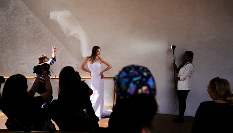 Woman in white dress posing on stage during a fashion or bridal show while photographers and spectators watch from the audience.