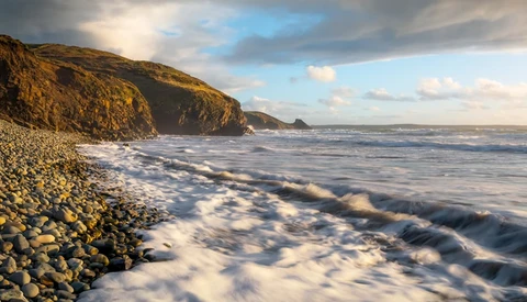Rocky coastline with steep cliffs and turbulent waves rolling onto a pebble beach at golden hour.