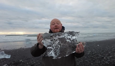 Man holding a large chunk of ice on a black sand beach with ocean and cloudy sky behind him.