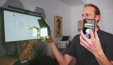 Photographer holding smartphone displaying color picker app while pointing at LED light positioned near computer monitor.