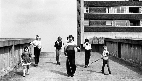 Children playing with a large tire on a rooftop concrete platform beside a brutalist apartment building.