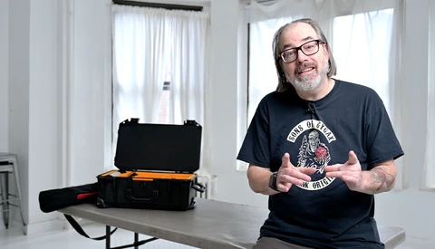 Man in glasses and band t-shirt gestures while speaking next to an open black and orange equipment case.