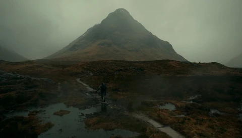 Lone hiker stands in wet moorland beneath a dramatic pyramidal mountain shrouded in mist.