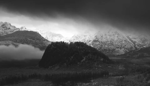 Snow-covered alpine peaks rise behind a forested ridge shrouded in mist under dark storm clouds.