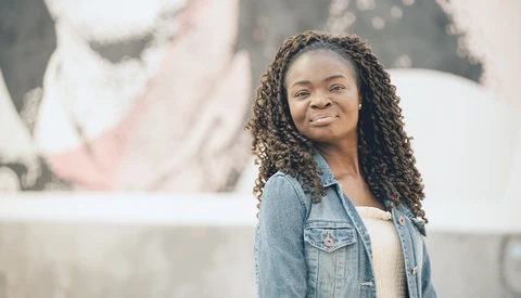 Woman with long braided hair wearing a denim shirt smiles at camera against blurred urban background.
