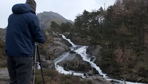 Photographer on tripod capturing a cascading mountain stream surrounded by evergreen forest and rocky terrain.