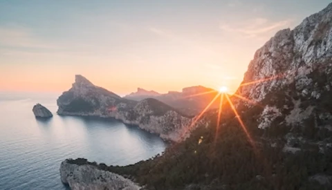 Sun bursting through rocky cliffs above a calm Mediterranean cove at sunrise.