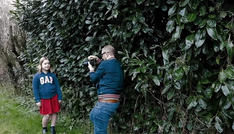Photographer taking a portrait of a young girl standing against dense ivy-covered foliage.