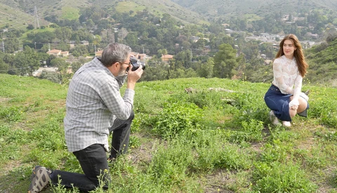 Photographer crouching while shooting a model posing in a grassy hillside field with forested mountains in the background.