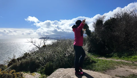 Person in magenta jacket photographing a coastal landscape with binoculars or telephoto lens.