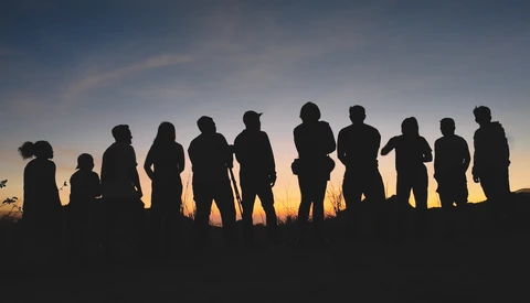 Silhouettes of approximately twelve people standing in a line against a golden sunset sky.
