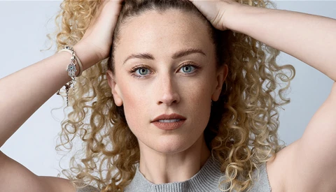 Woman with voluminous curly blonde hair posing with hands near head against neutral background.