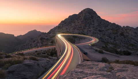 Long exposure light trails curve through a desert mountain pass at sunset.