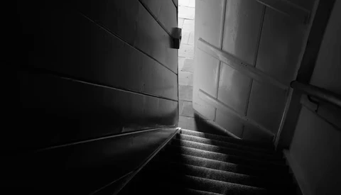 Narrow stairwell with stark contrast between dark and light areas, steps descending toward a bright doorway.
