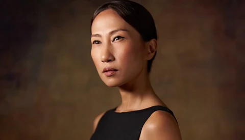 Woman in black sleeveless top against warm, blurred brown background, gazing to the side.