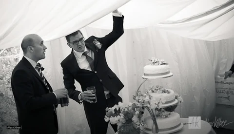 Two men in formal attire gesture toward a tiered wedding cake in a white draped tent setting.