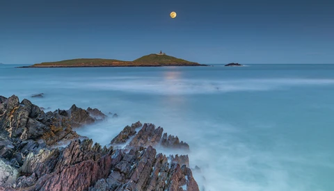 Rocky coastline with smooth water and distant island under clear sky with moon.