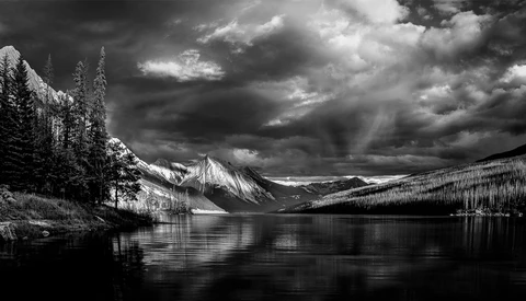 Snow-capped mountain reflected in still lake surrounded by evergreen forest under dramatic storm clouds.