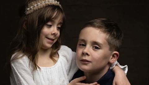Young girl with tiara embracing boy from behind against dark background.