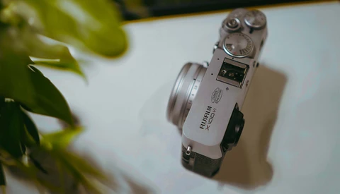Silver rangefinder camera with dual rewind knobs viewed from above on white surface with green plant leaf in frame.