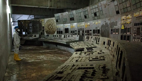 Abandoned control room with operator station and wall-mounted instrument panels covered in dust and decay.