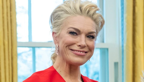 Woman with blonde hair styled in a braid, wearing a red top, smiling at camera in a bright interior space.
