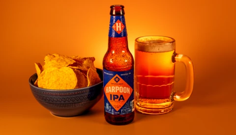 Bottle of IPA beer with glass mug and bowl of potato chips against warm orange background.