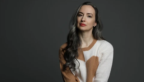 Woman with long dark hair wearing a cream and tan ribbed knit sweater against a gray background.