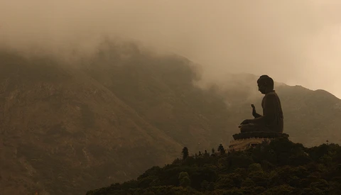 Large Buddha statue silhouetted against misty mountains shrouded in fog and clouds.