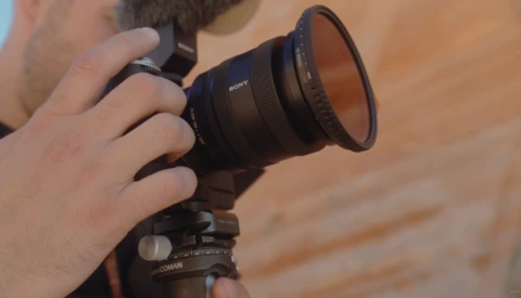Photographer holding a Sony telephoto lens mounted on a tripod with ball head.