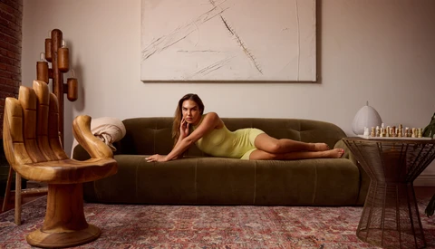 Woman in yellow dress reclines on olive green sofa in styled interior with sculptural wooden furniture.